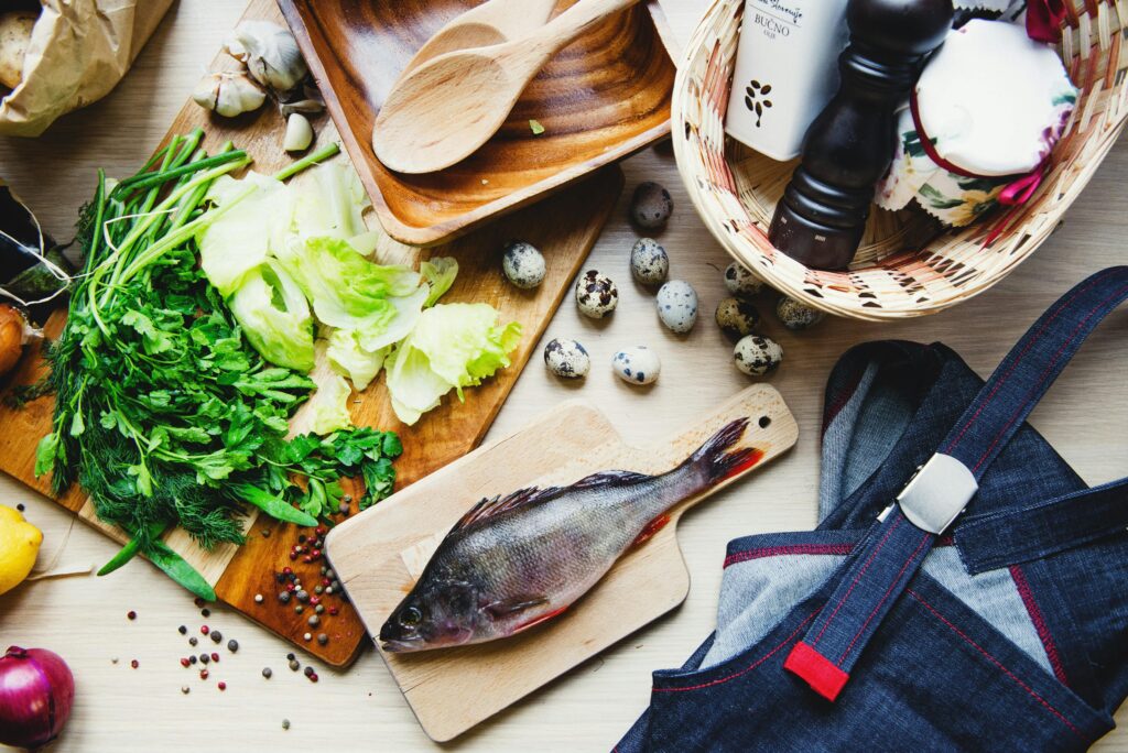 service-06 Top view of fresh fish and vegetables put on cutting board near wooden dishware and wicker basket with pepper shaker and jar in kitchen